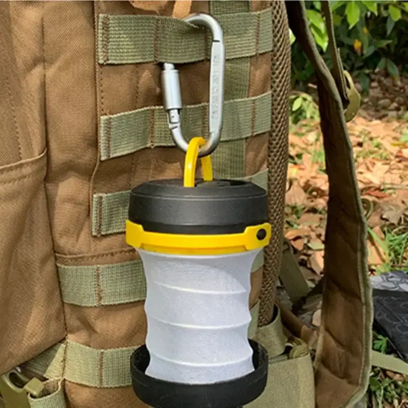 Collapsible camping lantern with yellow and black accents attached to a backpack strap.