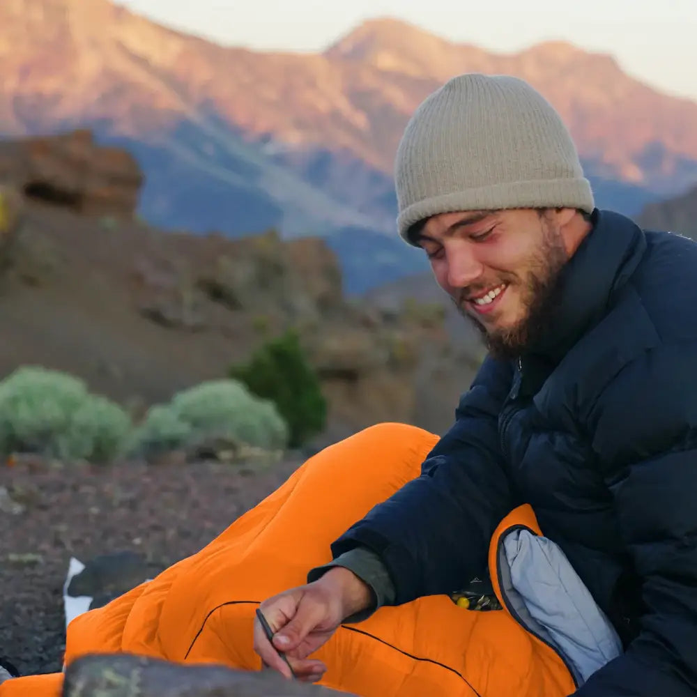 Smiling man in a beanie and jacket setting up an orange tent outdoors.