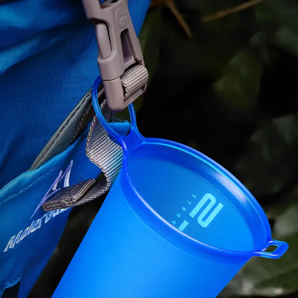 Blue collapsible silicone camping cup with measurement markings attached to a backpack strap.